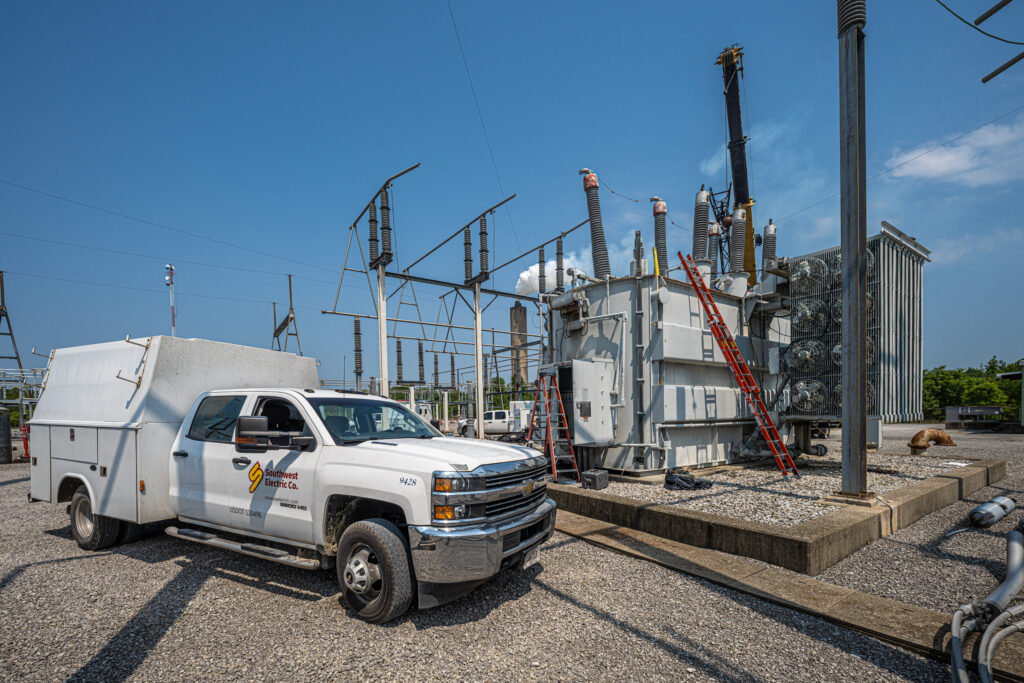 Utility maintenance truck near a high-voltage transformer in a substation, with ladders and cooling fans visible, indicating ongoing maintenance work under a clear blue sky.